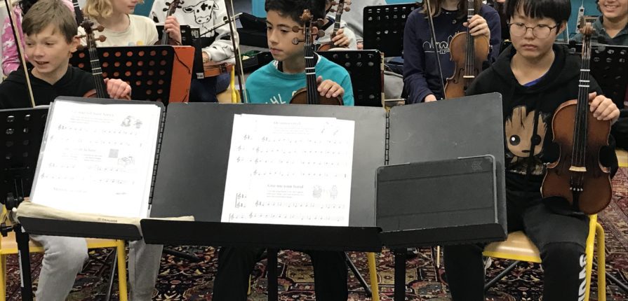 Students sitting in front of sheet music holding string instruments