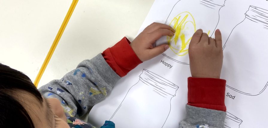 A kindergarten student colouring a jar with the word "Happy" below it to describe their feelings.