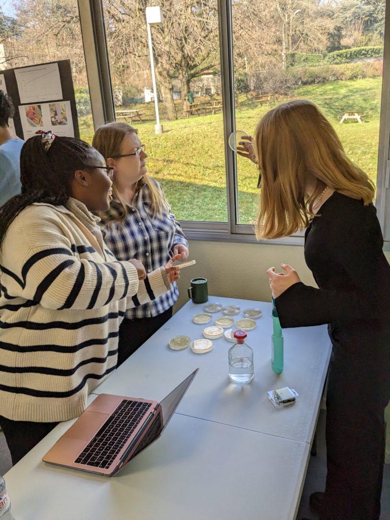 A student holding a petri dish up to a window for a teacher and another student to examine