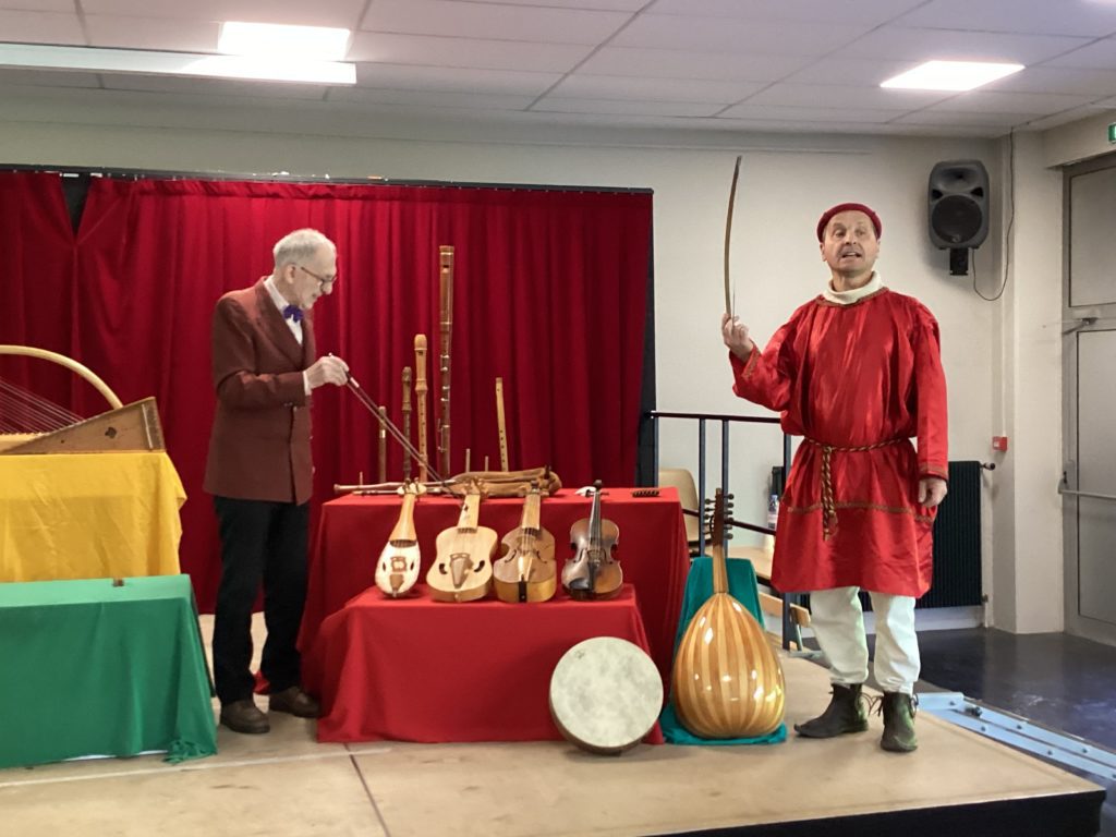 Two men on a stage, one dressed in medieval clothing, with period musical instruments