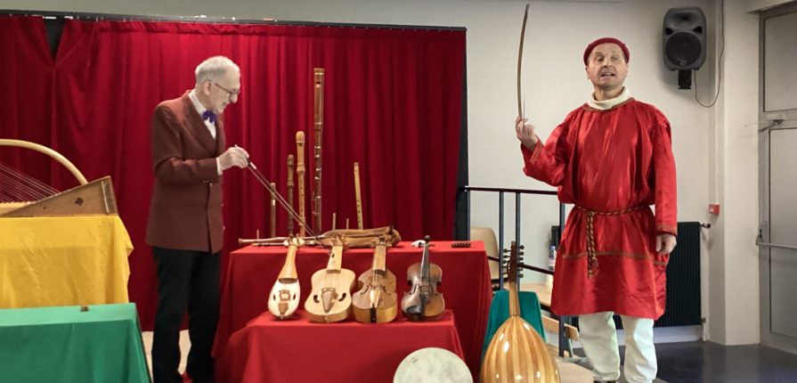 Two men on a stage, one dressed in medieval clothing, with period musical instruments