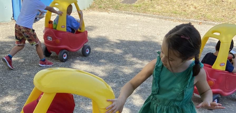 Kindergarten students driving toy cars during their outdoor learning lessons