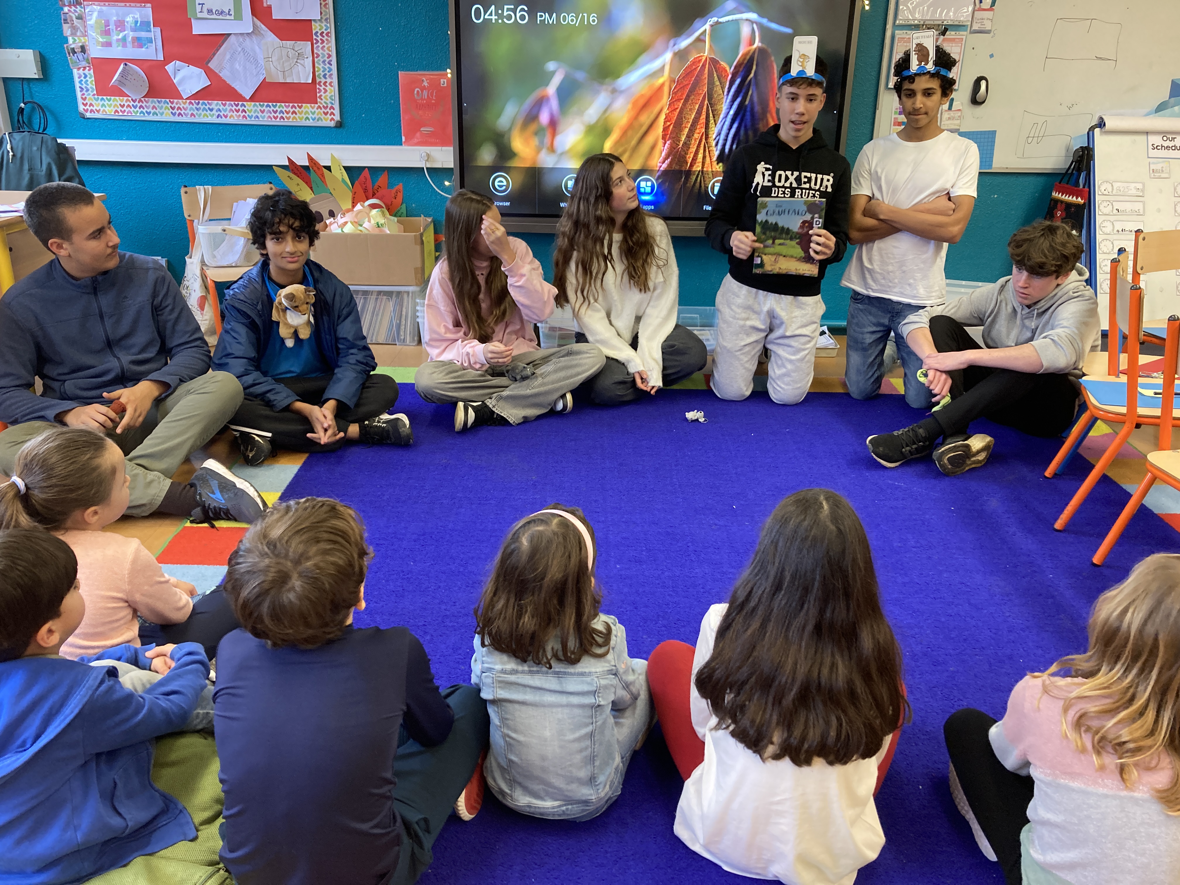 Grade 9 and kindergarten students sitting in a circle on a blue carpet in a kindergarten classroom.