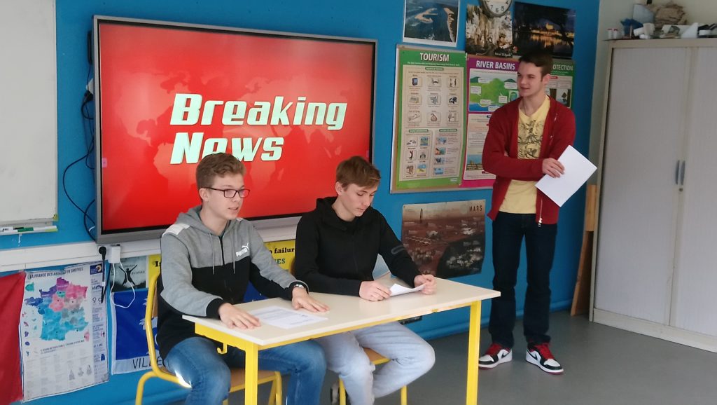 2 students seated in front of a screen with "Breaking News" written on it, 1 other student is standing next to the screen.