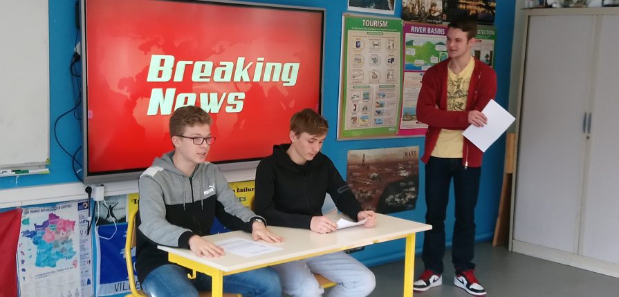 2 students seated in front of a screen with "Breaking News" written on it, 1 other student is standing next to the screen.