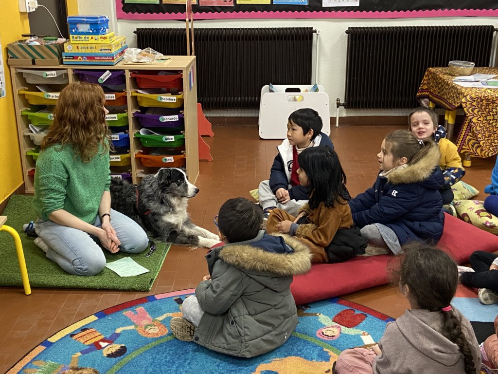 Students sitting on colourful carpets, looking at a dog and her owner