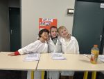 Three Grade 1 and 2 students wearing lab coats and posing together in front of desks