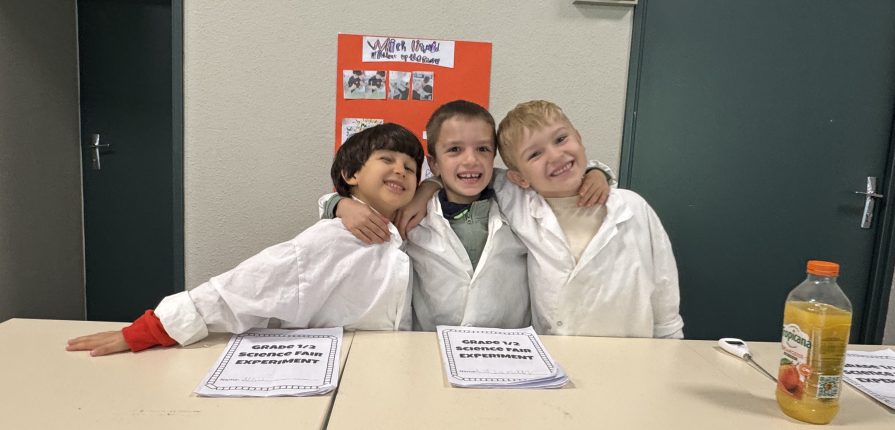Three Grade 1 and 2 students wearing lab coats and posing together in front of desks